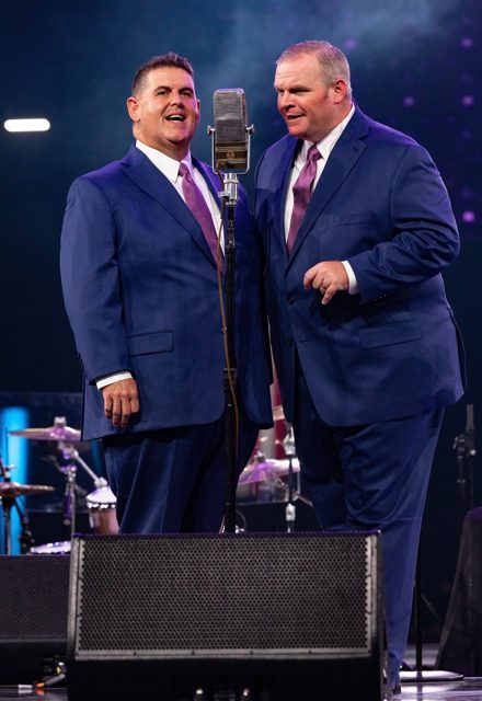 Two members of the Wills Brothers Quartet singing around a vintage microphone on the NQC MainStage